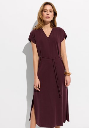 Kobieta o jasnobrązowych włosach, ubrana w bordową sukienkę midi z krótkim rękawem, dekoltem w serek, paskiem w talii i bocznymi rozcięciami, stojąca na białym tle.