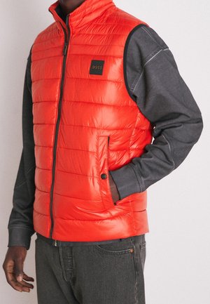 Mężczyzna ubrany w jaskrawo pomarańczową pikowaną kamizelkę na ciemnoszarym długim rękawie oraz ciemne dżinsy, z ręką w kieszeni kamizelki, stojący na jednolitym tle.