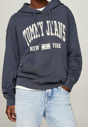Donkerblauwe hoodie met de tekst en het logo "Tommy Jeans New York", gedragen met lichtblauwe spijkerbroek, hand van persoon in zak gestoken.