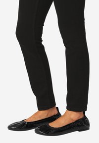 Stopy w czarnych, błyszczących balerinach z wzorem wężowej skóry, zestawione z wąskimi czarnymi spodniami na białym tle.