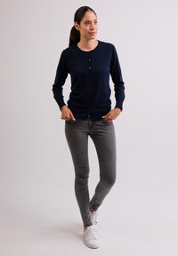 Námorně modrý svetr s kulatým výstřihem, zapínáním na knoflíky a dlouhými rukávy, spárovaný s úzkými šedými džíny a bílými teniskami. Jednoduchý design.