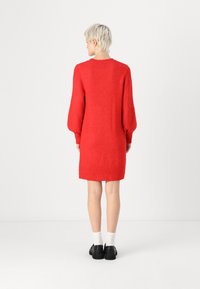 Robe tricotée rouge avec de longues manches bouffantes, un col rond et des poignets côtelés. Le tissu semble doux et texturé. Chaussures noires et chaussettes blanches.
