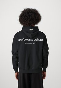 Schwarzer Hoodie, von hinten angezeigt, mit weißem Text "don't waste culture" und "MAISON D'ART." Enthält eine passgenaue Kapuze und gerippte Bündchen.
