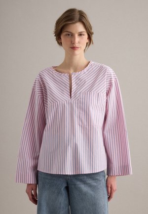 Joven mujer con el cabello corto que lleva una camisa rosa de manga larga con rayas verticales azules y vaqueros azul claro, de pie frente a un fondo liso.