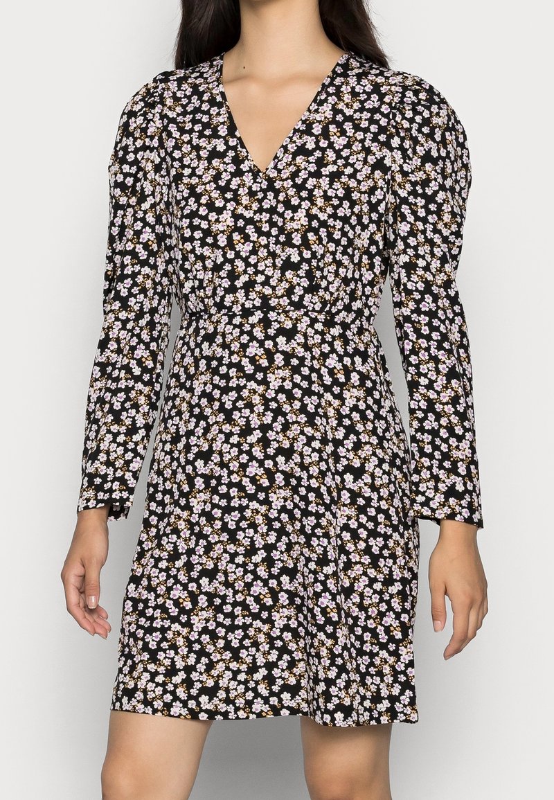 Femme portant une robe noire à manches longues avec un petit motif floral blanc et violet clair, décolleté en V et jupe légèrement évasée.