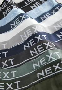 Daudzkrāsains stiepjamais audums ar joslām, kurās attēlota "NEXT" logo baltā krāsā. Krāsas ietver melnu, pelēku, tumši zilu, gaiši zilu un zaļu.