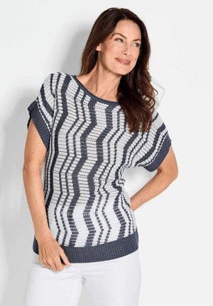 Strickpullover mit Zickzackmuster in Marineblau und Weiß, mit kurzen Ärmeln, einem runden Ausschnitt und einem gerippten Saum, getragen mit weißen Hosen.