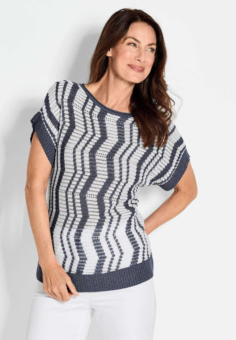 Strickpullover mit Zickzackmuster in Marineblau und Weiß, mit kurzen Ärmeln, einem runden Ausschnitt und einem gerippten Saum, getragen mit weißen Hosen.
