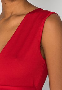 Robe rouge sans manches avec un décolleté en V profond et un tissu lisse et extensible. Le design structuré met en valeur les épaules et la poitrine.