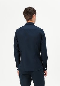 Camicia blu navy con bottoni, realizzata in tessuto liscio, dotata di colletto, maniche lunghe e orlo arrotondato sul retro, con dettagli in bianco sui bottoni.