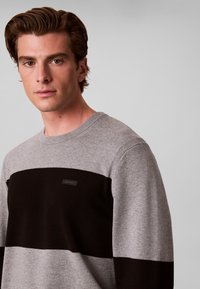 Jeune homme aux cheveux courts et bruns portant un pull bicolore gris et noir, regardant légèrement vers la caméra sur un fond clair uni.