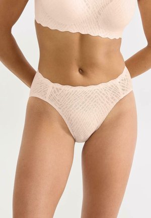 Kobieta ubrana w jasnobeżową, wzorzystą bieliznę z wysokim stanem oraz dopasowany biustonosz, stojąca z rękami za plecami na jednolitym tle.