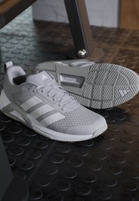 Paire de baskets Adidas grises sur un sol de gym noir texturé, une chaussure debout et l'autre montrant la semelle avec le logo Adidas.