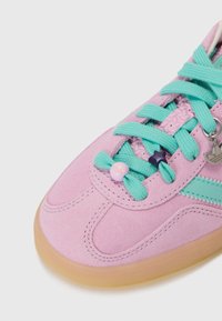Sneakers en suède rose clair avec des lacets turquoise, une bande aqua et des charms décoratifs, dotés d'une semelle en gomme flexible et de détails texturés.
