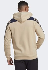Hombre con sudadera beige y azul marino con tres rayas en los hombros, de pie de espaldas a la cámara contra un fondo liso.