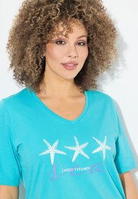 T-shirt turchese a V con tre grafiche di stelle marine bianche e il testo "HO BISOGNO DI VITAMINA MARE" in viola, realizzato in morbido cotone.