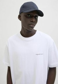 T-shirt en coton blanc avec un petit logo brodé "originals studio". Le modèle porte une casquette bleu marine en tissu texturé.