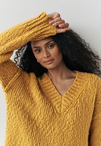 Chunky strikket sweater i sennepsgul, med V-hals design, ribbet manchetter og tekstureret stof. Varm og oversized pasform.