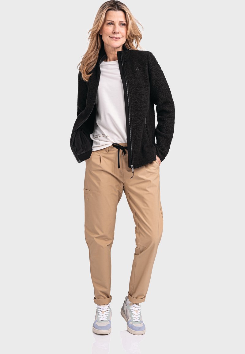 Giacca con zip in pile nera, camicia bianca, pantaloni beige con vita elasticizzata e scarpe da ginnastica azzurre chiare. L'outfit presenta una vestibilità rilassata e casual.
