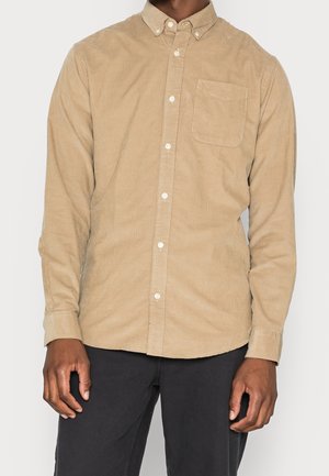 Homme portant une chemise beige en velours côtelé à boutons avec une poche avant et un pantalon noir sur un fond blanc uni.