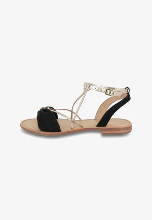 Sandali in suede nero e dettagli metallici dorati con un design a strisce, suola piatta e cinturino alla caviglia. Presentano una punta arrotondata e un tallone aperto.