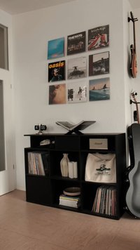 Zwart houten kast met kubusvormige opbergruimte, vinylplaten, decoratieve items en een zilveren klok. Aan de muur hangen ingelijste albumhoezen.