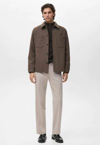Camicia giacca marrone in misto lana con collo a camicia, due tasche anteriori, indossata sopra un maglione lavorato a maglia scuro e pantaloni beige chiaro.