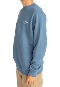 Sudadera de cuello redondo azul, hecha de una mezcla de algodón, con puños y cintura acanalados y un logotipo en texto blanco en el pecho. Textura suave.