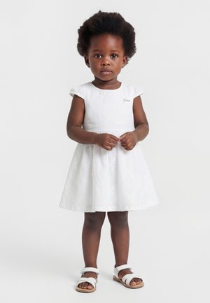 Petite fille aux cheveux bouclés portant une robe blanche à manches courtes et des sandales blanches, debout devant un fond uni.