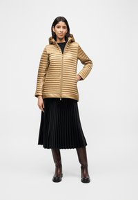 Γυναίκα που φορά χρυσό μπουφάν με καπιτονέ σχέδιο, μαύρη πλισέ μίντι φούστα και σκούρα καφέ μπότες μέχρι το γόνατο, στέκεται μπροστά σε ένα απλό φωτεινό φόντο.