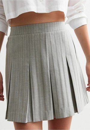 Femme portant une jupe plissée à taille haute grise à fines rayures, associée à une chemise blanche courte aux manches retroussées.