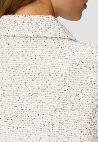 Strukturiertes weißes Tweed-Gewebe mit schwarzen Sprenkeln. Der Kragen ist klassisch und strukturiert und zeigt ein gewebtes Muster sowie subtile Details.