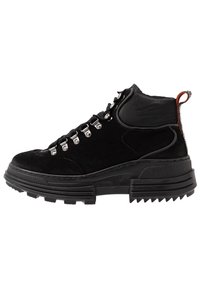 Bota negra de media caña confeccionada en suede y goma, con ojales metálicos, suela gruesa con tacos y un tirador en el talón.