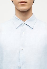 Młoda osoba ubrana w jasnoniebieską koszulę zapinaną na guziki z klasycznym kołnierzykiem na jednolitym białym tle.