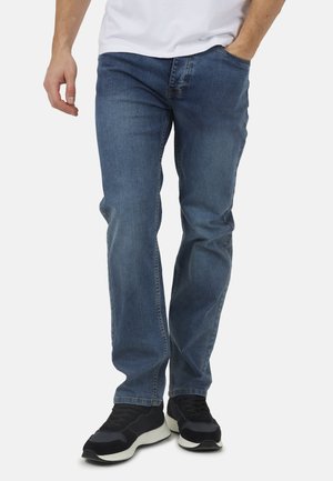 Uomo con jeans di denim blu e scarpe da ginnastica nere, in piedi con una mano in tasca contro uno sfondo semplice.