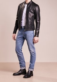 Man som bär en svart läderjacka, vit skjorta, ljusblå uppvikta jeans och svarta läderstövlar som står på en beige golv.