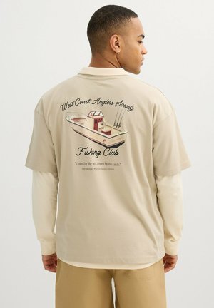 Hombre con camisa beige de capas con gráfico de barco de pesca y texto "West Coast Anglers Society Fishing Club" en la parte trasera