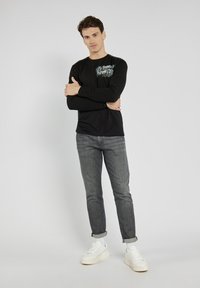 Czarna bluzka z długim rękawem z graficznym nadrukiem na górnej lewej części; w połączeniu z szarymi dopasowanymi dżinsami i białymi sneakersami. Gładka faktura materiału.