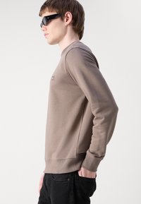 Sudadera marrón claro con mangas largas, puños acanalados y un pequeño logo en el pecho, combinada con pantalones negros y gafas de sol oscuras.