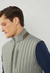 Tkanina siva prsluk s quilted dizajnom i visokim ovratnikom, s džepom na prsima i smešnim akcentom na ramenu. Nosi se preko tamnoplave košulje dugih rukava.