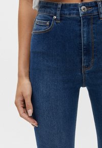 Modré džínové kalhoty s vysokým pasem a hladkou texturou, vybavené přední kapsou, zapínáním na knoflík a kontrastním oranžovým prošitím.