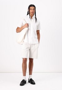 Witte kortemouwen blouse, gestreepte crèmekleurige shorts, zwarte loafers en een crèmekleurige tote bag. Model staat tegen een effen achtergrond.