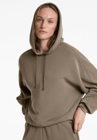 Oversized olive green hoodie in soft fabric with a drawstring hood, ribbed accents on sleeves, and relaxed fit. Styled with matching pants.