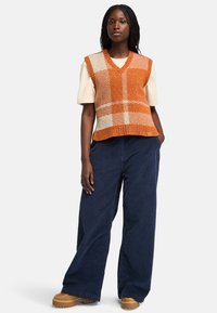 Chaleco de punto en naranja y crema con escote en V, que presenta un patrón a cuadros. Combinado con pantalones de pana anchos en color marino y zapatos de plataforma marrones.