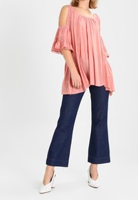 Růžová top bez ramínek s volnými rukávy a detailním prošíváním, zkombinovaný s tmavě modrými volnými džínami a bílými lodičkami na vysokém podpatku.