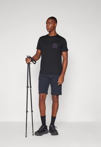 Svart t-skjorte med korte ermer og et sirkulært lilla logo, kombinert med marineblå shorts. Personen holder turseskaft og har på seg svarte joggesko.