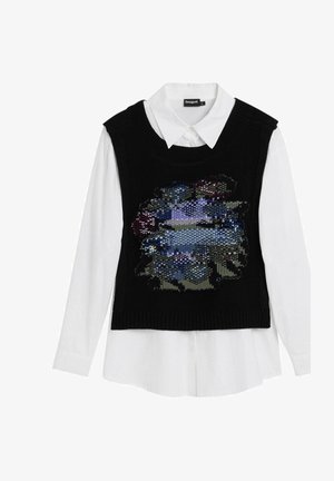 Vesta din tricot cu un design abstract colorat din mărgele, de culoare neagră, purtată peste o cămașă albă cu guler. Mâneci lungi vizibile.