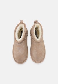 Bottines couleur noisette en daim doux avec une doublure en fausse fourrure blanche et moelleuse. Bout arrondi, texture lisse, et languettes à l'arrière.