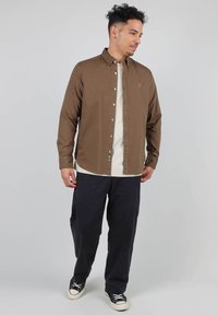 Chemise boutonnée à manches longues marron par-dessus un t-shirt crème, associée à un pantalon bleu marine foncé et des baskets noir et blanc.