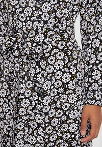 Tessuto nero con motivo floreale bianco e giallo; presenta una vita annodata, maniche lunghe e design con bottoni. Dettaglio del tessuto testurizzato visibile.